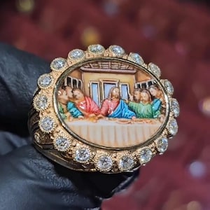 Può includere: Anello d'oro con una miniatura dell'Ultima Cena. Il centro ovale è circondato da piccoli diamanti scintillanti. L'anello ha una fascia spessa. Il dipinto raffigura figure in abiti colorati.