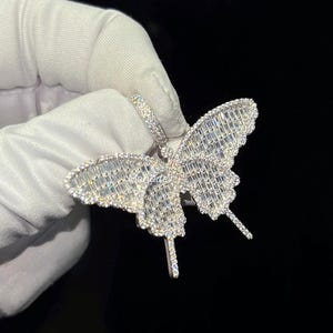 May include: A silver butterfly pendant with a sparkling, gem-studded design. The pendant features a detailed butterfly shape with intricate patterns and a loop for a chain. The wings are filled with rectangular gems.
