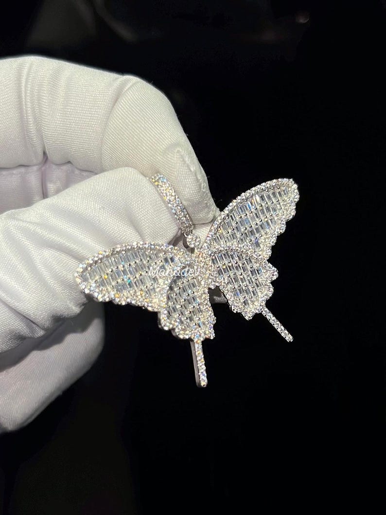 May include: A silver butterfly pendant with a sparkling, gem-studded design. The pendant features a detailed butterfly shape with intricate patterns and a loop for a chain. The wings are filled with rectangular gems.