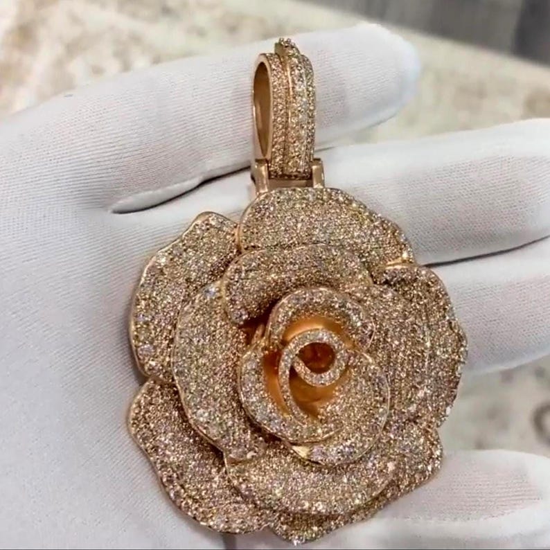 May include: Rose-shaped pendant with a rose gold finish, encrusted with numerous small, sparkling stones. The pendant has a bail for attaching to a chain. The jewelry is held by a gloved hand.