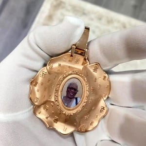 May include: A gold-colored pendant in the shape of a flower, featuring an oval photo of a person. The pendant has a decorative border and is held by a gloved hand. The flower petals have a textured design.