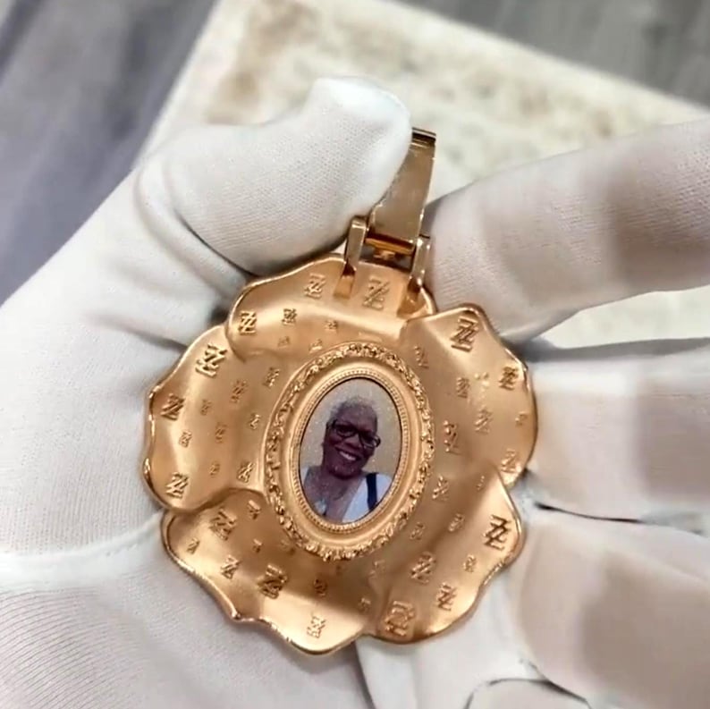 May include: A gold-colored pendant in the shape of a flower, featuring an oval photo of a person. The pendant has a decorative border and is held by a gloved hand. The flower petals have a textured design.