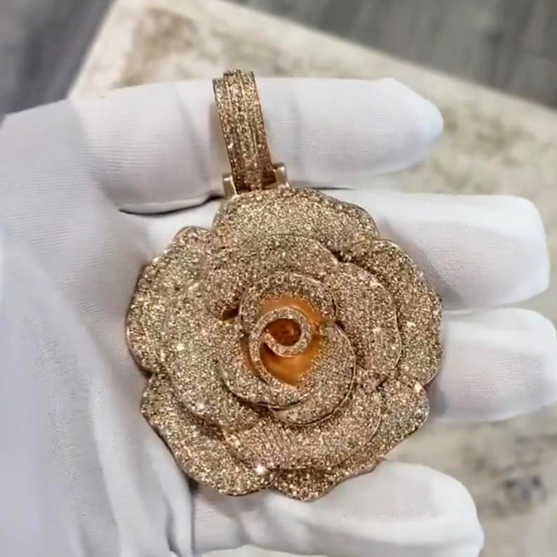 May include: A rose-shaped pendant with a gold-colored finish, encrusted with numerous small, sparkling stones. The pendant has a loop at the top for a chain. The rose's petals are detailed, and the center is a lighter gold color.