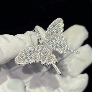 May include: A silver butterfly pendant encrusted with sparkling, clear gemstones. The butterfly design features detailed wings and antennae. The pendant is held against a white background.
