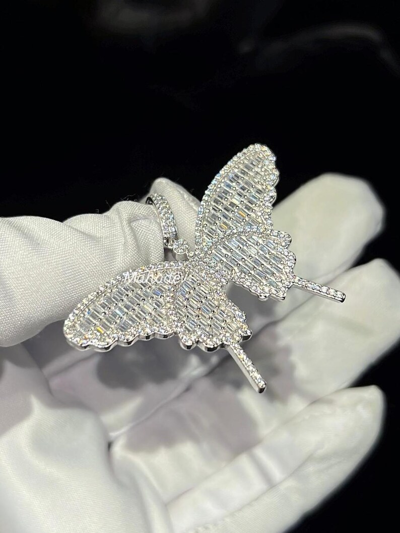 May include: A silver butterfly pendant encrusted with sparkling, clear gemstones. The butterfly design features detailed wings and antennae. The pendant is held against a white background.