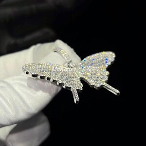 May include: A silver butterfly pendant encrusted with numerous small, clear gemstones. The pendant is highly detailed, with the wings and body of the butterfly clearly defined. The pendant is held against a dark background.