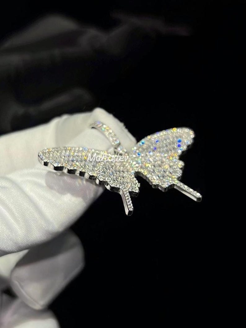 May include: A silver butterfly pendant encrusted with numerous small, clear gemstones. The pendant is highly detailed, with the wings and body of the butterfly clearly defined. The pendant is held against a dark background.