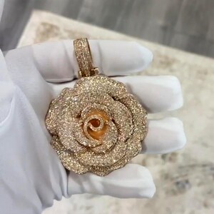 May include: A rose-shaped pendant with a sparkling, gold-colored finish. The pendant features a detailed design with multiple layers of petals and a loop for a chain. The surface is covered in small, glittering accents, giving it a luxurious appearance.