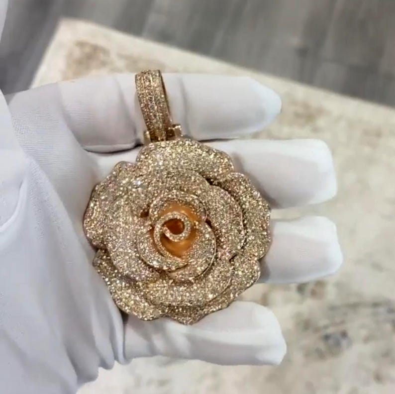 May include: A rose-shaped pendant with a sparkling, gold-colored finish. The pendant features a detailed design with multiple layers of petals and a loop for a chain. The surface is covered in small, glittering accents, giving it a luxurious appearance.