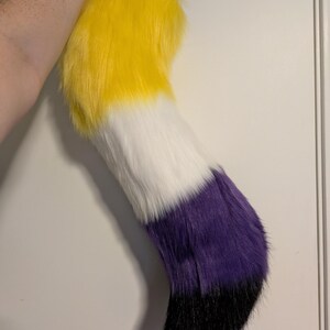 May include: A plush, faux fur tail in the non-binary pride flag colors: yellow, white, purple, and black. The tail is held up against a white background. The tail is a fun accessory for cosplay or costume events.