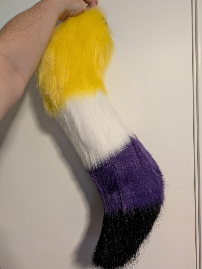 May include: A plush, faux fur tail in the non-binary pride flag colors: yellow, white, purple, and black. The tail is held up against a white background. The tail is a fun accessory for cosplay or costume events.