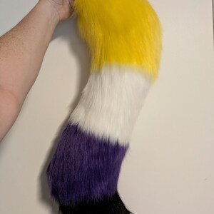May include: A plush, furry tail in the colors of yellow, white, purple, and black. The tail is held up by a hand, showing the top portion of the tail in yellow, transitioning to white, then purple, and ending in black.