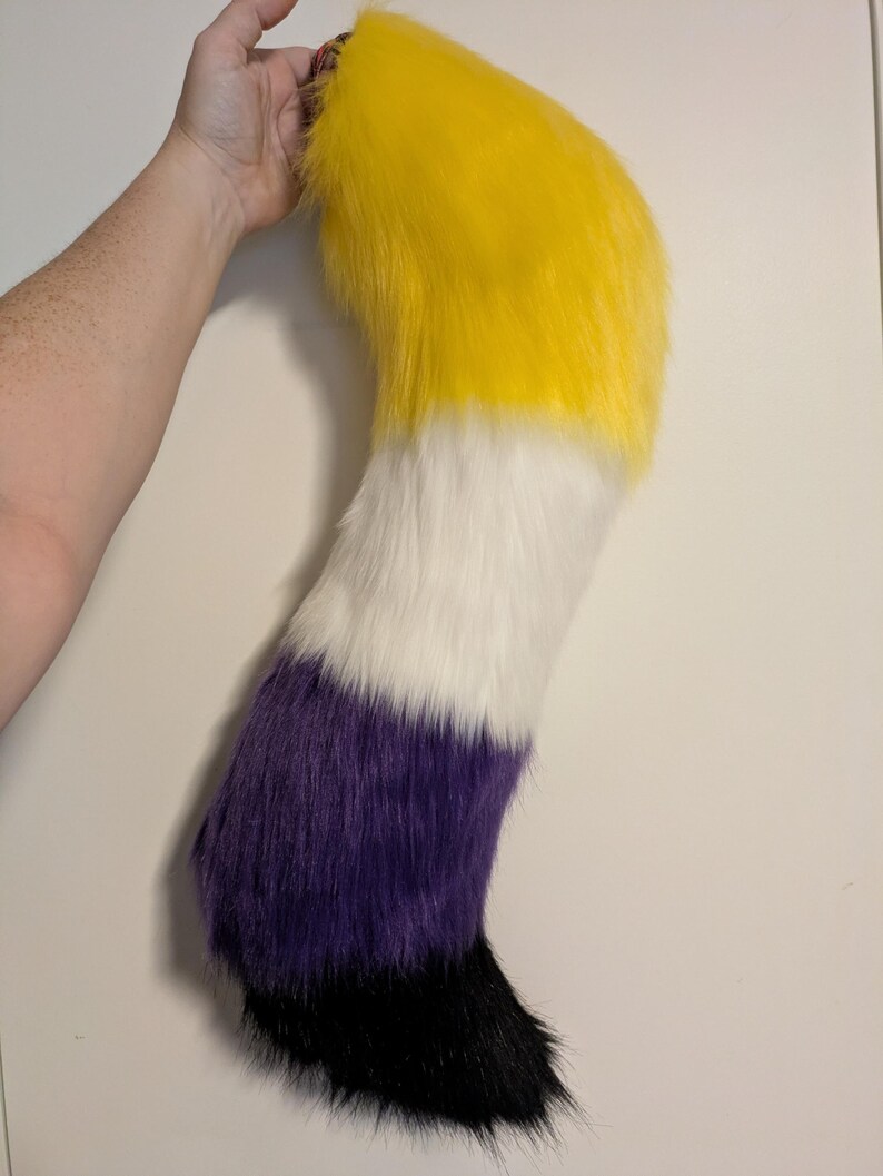 May include: A plush, furry tail in the colors of yellow, white, purple, and black. The tail is held up by a hand, showing the top portion of the tail in yellow, transitioning to white, then purple, and ending in black.
