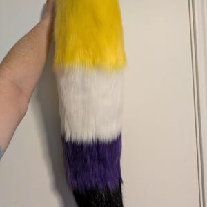 May include: A faux fur tail in the non-binary pride flag colors: yellow, white, purple, and black. The tail is held up against a white background. The tail is made of soft, fluffy material.