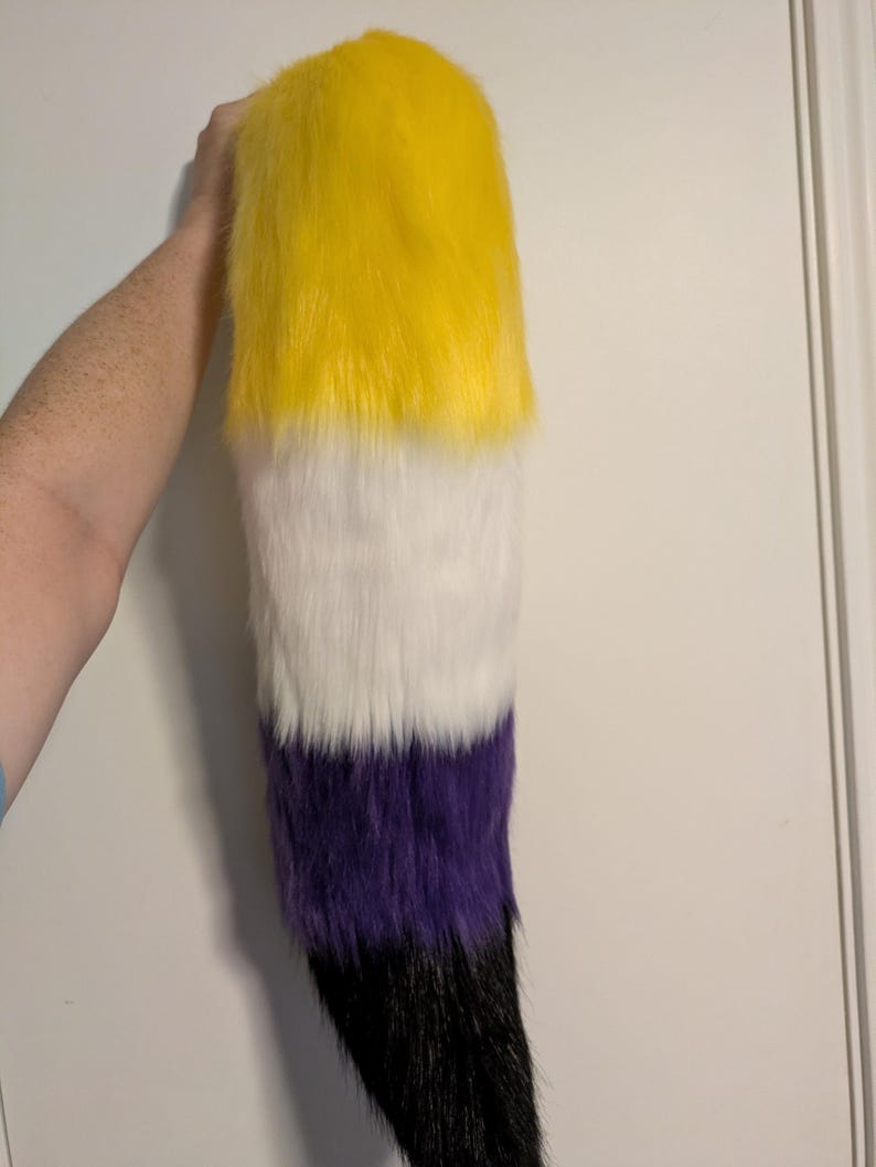 May include: A faux fur tail in the non-binary pride flag colors: yellow, white, purple, and black. The tail is held up against a white background. The tail is made of soft, fluffy material.