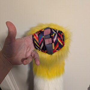May include: A yellow, white, and purple faux fur tail with a colorful patterned wrist strap. The tail is yellow at the top, fading to white, and ending in purple. The wrist strap has a geometric design.