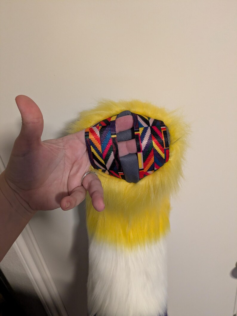 May include: A yellow, white, and purple faux fur tail with a colorful patterned wrist strap. The tail is yellow at the top, fading to white, and ending in purple. The wrist strap has a geometric design.