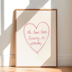 Op de afbeelding: Een ingelijste print met een lichte achtergrond en een houten frame. De print toont een roze hart met de handgeschreven tekst, "oh, how I love being a woman."
