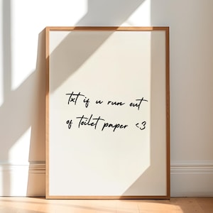 May include: A framed print with the text "Txt if u run out of toilet paper <3" in black script. The print has a cream-colored background and a wooden frame. The print is leaning against a white wall on a wooden floor.