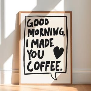 May include: A framed art print with the text "GOOD MORNING, I MADE YOU COFFEE" in black on a cream background. The print includes a black heart and is set in a light brown wooden frame. The artwork is displayed on a wooden floor.