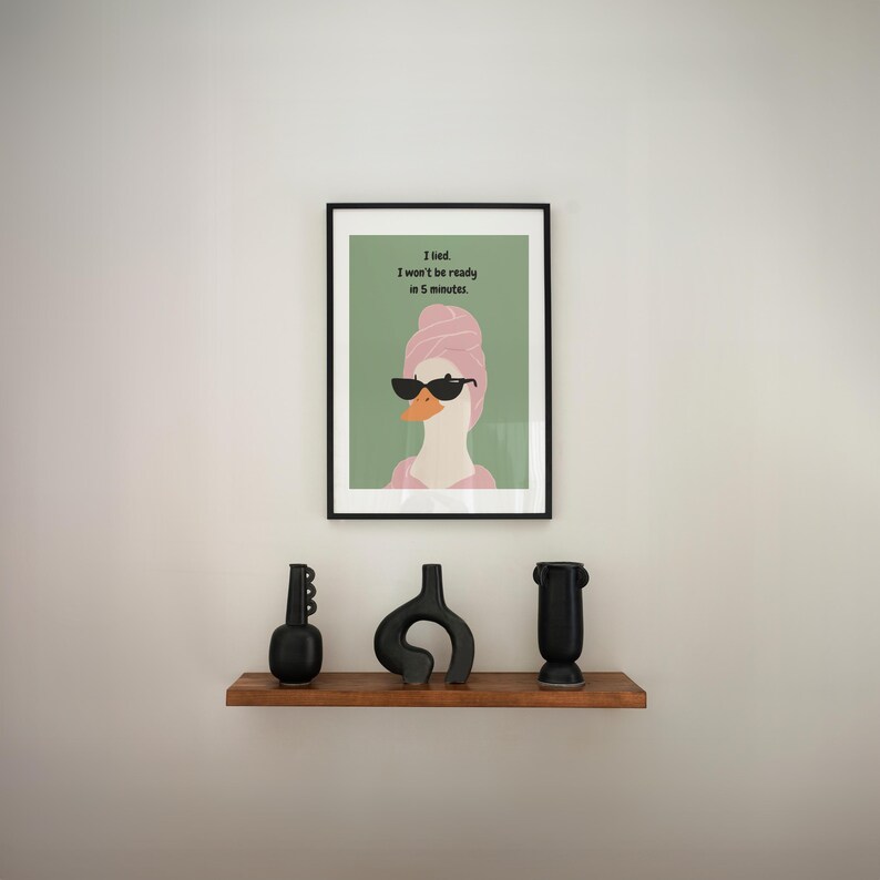 Puede incluir: Una impresi&oacute;n art&iacute;stica enmarcada con un ganso con gafas de sol y una toalla rosa, con el texto "I lied. I won't be ready in 5 minutes." Debajo, una estanter&iacute;a de madera exhibe tres jarrones de cer&aacute;mica negros.