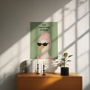 Puede incluir: Un cuadro enmarcado con un pato de dibujos animados con gafas de sol y una toalla rosa en la cabeza. El cuadro tiene el texto "I lied. I won't be ready in 5 minutes". La obra de arte se exhibe en una pared.
