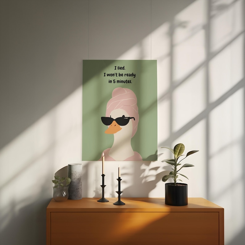 Puede incluir: Un cuadro enmarcado con un pato de dibujos animados con gafas de sol y una toalla rosa en la cabeza. El cuadro tiene el texto "I lied. I won't be ready in 5 minutes". La obra de arte se exhibe en una pared.