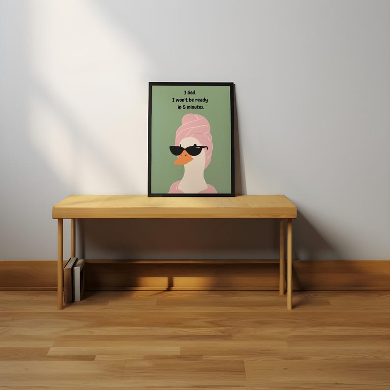 Puede incluir: Una impresi&oacute;n art&iacute;stica enmarcada que presenta un ganso de dibujos animados con gafas de sol y una toalla rosa en la cabeza. La impresi&oacute;n tiene el texto: "I lied. I won't be ready in 5 minutes." La obra est&aacute; sobre un banco de madera.