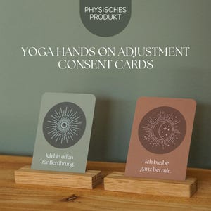 Könnte beinhalten: Zwei Karten mit Himmelsmotiven, die auf Holzblöcken präsentiert werden. Auf den Karten steht "YOGA HANDS ON ADJUSTMENT CONSENT CARDS" und sie enthalten deutschen Text. Eine Karte ist salbeigrün mit einem Sonnenstrahl-Design, die andere ist terrakottafarben mit Mond und Sternen.