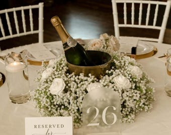 May include: A champagne bottle in a gold metal bucket surrounded by a wreath of white roses and baby's breath. An arched acrylic table number sign displays the number 26. A white card reads RESERVED for FAMILY. A glass candle holder is nearby.