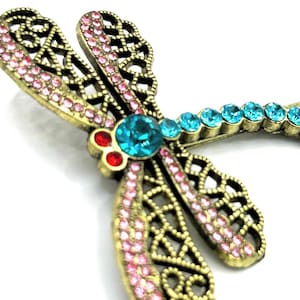 May include: A detailed close-up of a dragonfly brooch. The brooch features a gold-toned metal frame with intricate cut-out designs. The wings are adorned with pink rhinestones, while the body has turquoise and red gemstones.