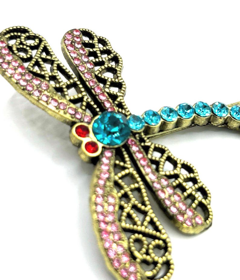 May include: A detailed close-up of a dragonfly brooch. The brooch features a gold-toned metal frame with intricate cut-out designs. The wings are adorned with pink rhinestones, while the body has turquoise and red gemstones.