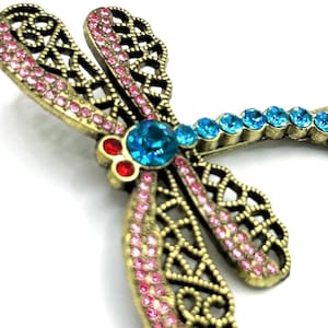 May include: A detailed dragonfly brooch with a bronze-colored metal frame. The wings are adorned with pink rhinestones, and the body features a large turquoise center stone, with smaller blue stones along the tail. Red stones accent the head.