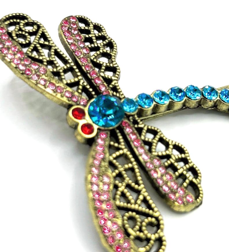 May include: A detailed dragonfly brooch with a bronze-colored metal frame. The wings are adorned with pink rhinestones, and the body features a large turquoise center stone, with smaller blue stones along the tail. Red stones accent the head.