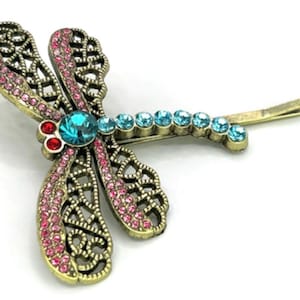 May include: A dragonfly hair clip with a bronze-colored metal frame. The wings are adorned with pink rhinestones, and the body features a large turquoise gemstone and smaller blue rhinestones. Red gemstones accent the head.