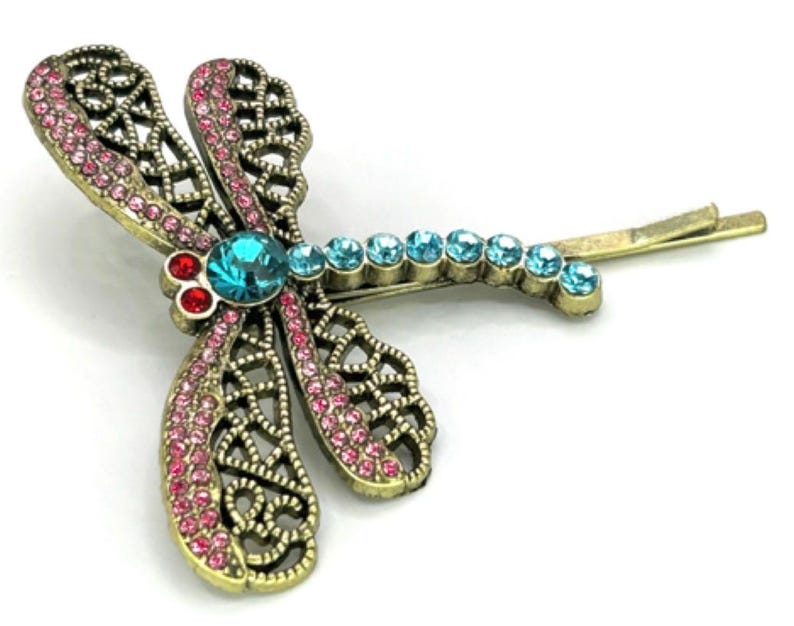May include: A dragonfly hair clip with a bronze-colored metal frame. The wings are adorned with pink rhinestones, and the body features a large turquoise gemstone and smaller blue rhinestones. Red gemstones accent the head.