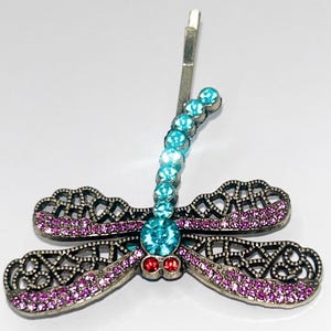 May include: A dragonfly hair clip with a silver-tone metal frame. The dragonfly's body is adorned with a row of light blue crystals, and the wings are embellished with pink crystals. Two red crystals are at the base of the body.