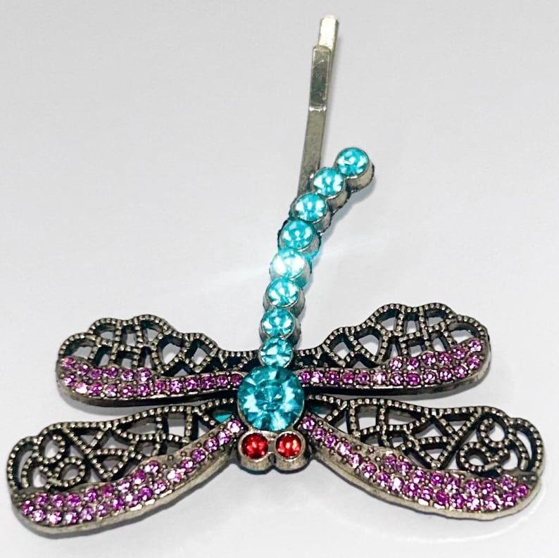 May include: A dragonfly hair clip with a silver-tone metal frame. The dragonfly's body is adorned with a row of light blue crystals, and the wings are embellished with pink crystals. Two red crystals are at the base of the body.