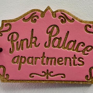 May include: A wooden sign with the words "Pink Palace apartments" carved into it. The sign has a pink background with gold-coloured trim and lettering. The sign is rectangular with a decorative top.