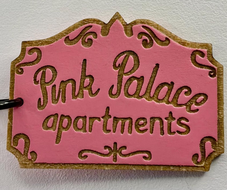 May include: A wooden sign with the words "Pink Palace apartments" carved into it. The sign has a pink background with gold-coloured trim and lettering. The sign is rectangular with a decorative top.