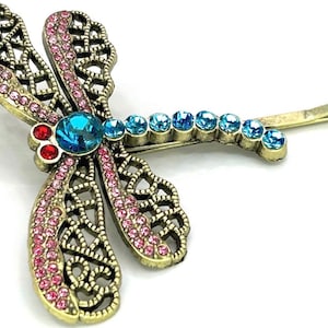 May include: A decorative dragonfly hair clip with a gold-tone metal frame. The wings are adorned with pink and blue rhinestones, and the body features a large blue rhinestone. The clip is designed to be inserted into the hair.