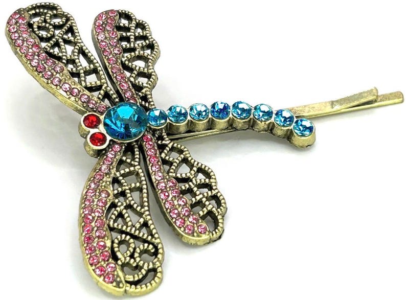 May include: A decorative dragonfly hair clip with a gold-tone metal frame. The wings are adorned with pink and blue rhinestones, and the body features a large blue rhinestone. The clip is designed to be inserted into the hair.