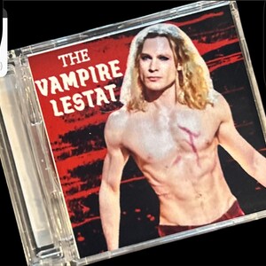 May include: A CD case featuring the text "THE VAMPIRE LESTAT" in white letters on a red background. The image shows a shirtless person with long blonde hair and a red mark on their chest.