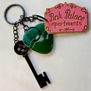 May include: A keychain with a black key, a green heart-shaped charm, and a pink sign reading "Pink Palace apartments" in gold. The keychain has a black ring and chain.