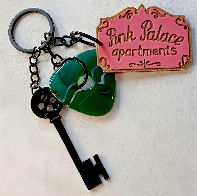 May include: A keychain with a black key, a green heart-shaped charm, and a pink sign reading "Pink Palace apartments" in gold. The keychain has a black ring and chain.