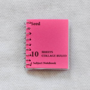 May include: A pink, 10-sheet, collage ruled subject notebook with a silver spiral binding. The notebook has the word "Seed" printed on the front cover.