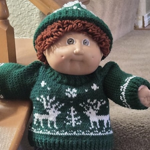 以下が含まれることがあります： 緑色のニットセーターに白いトナカイの模様、茶色のニット帽に白いポンポン、青いジーンズ、白いスニーカーを履いたキャベツパッチキッズ人形です。