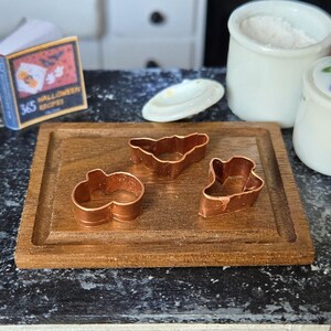Può includere: Tre stampini per biscotti in rame a forma di pipistrello, zucca e fantasma su un tagliere di legno. Gli stampini per biscotti sono su un piano di lavoro nero con due barattoli di ceramica bianca sullo sfondo.