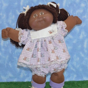 May include: A brown-skinned Cabbage Patch Kid doll with brown hair in pigtails wearing a lavender and white gingham dress with a white lace trim and white shoes.