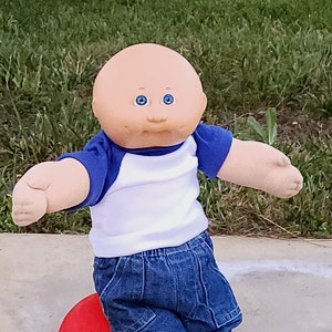 Può includere: Bambola Cabbage Patch Kid con una maglietta da baseball blu e bianca, jeans e scarpe da ginnastica bianche con strisce blu. La bambola è seduta su una palla rossa, con uno sfondo erboso.